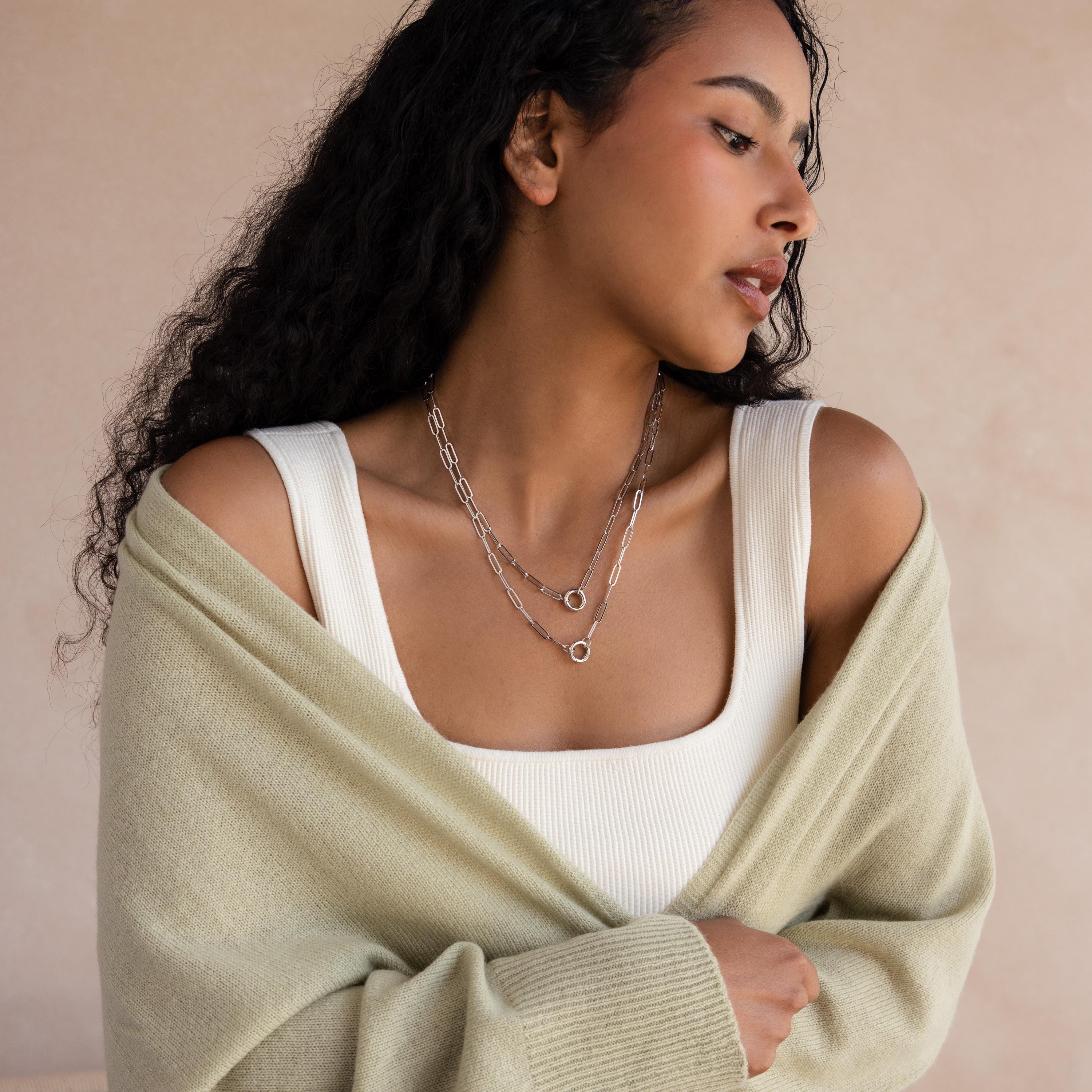 Woman with long curly hair wearing a Paperclip Clasp Necklace in White Gold, layered silver necklaces, a white top, and a light green cardigan.
