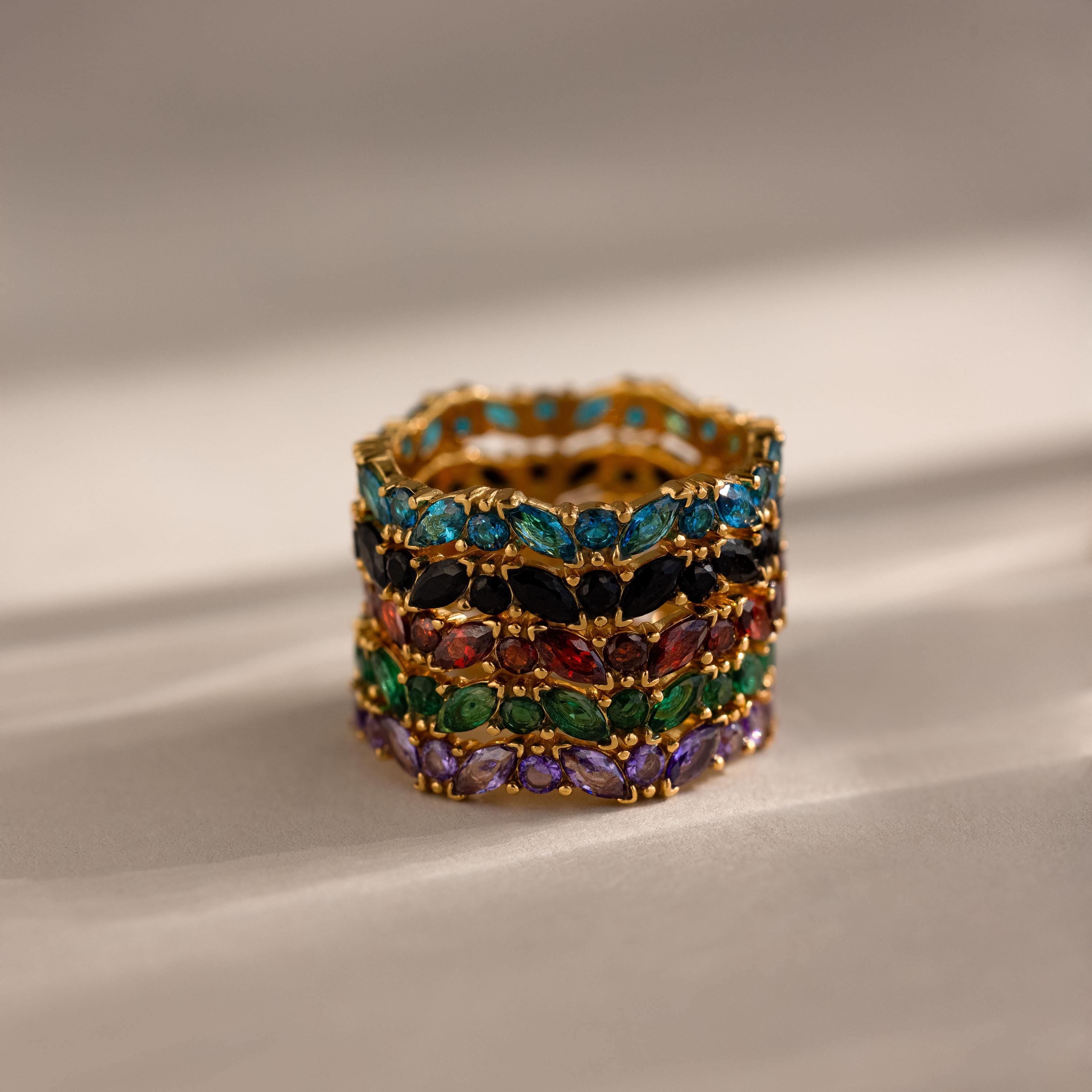 The Staggered Marquise Birthstone Ring, a gold band adorned with vibrant gemstones, is elegantly displayed on a neutral surface under gentle lighting.