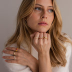 Woman with wavy blonde hair wears the Mixed Art Deco Birthstone Ring in 18K Gold, featuring red and clear stones, as she gazes thoughtfully to the side.