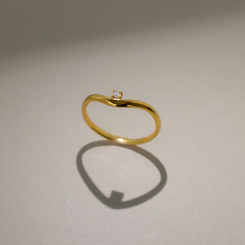 The Birthstone Drop Curve Ring, featuring a delicate gold band and a petite gem, casts a soft shadow on a beige surface.