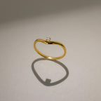 The Birthstone Drop Curve Ring, featuring a delicate gold band and a petite gem, casts a soft shadow on a beige surface.
