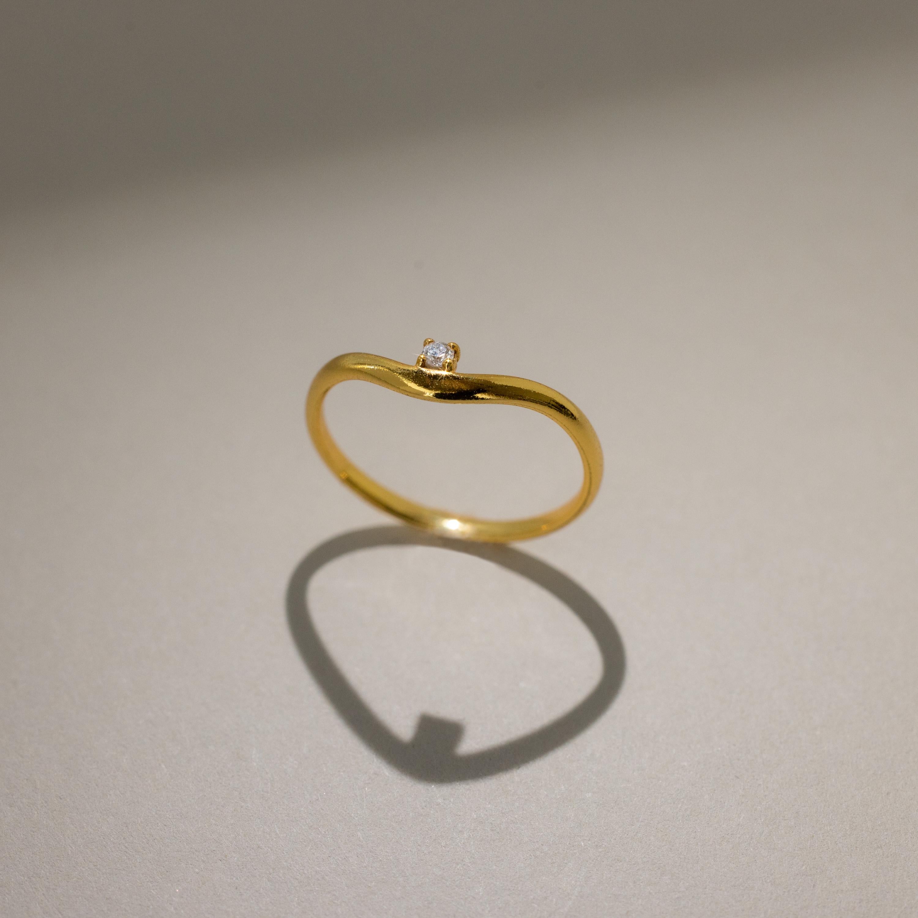 The Birthstone Drop Curve Ring, featuring a delicate gold band and a petite gem, casts a soft shadow on a beige surface.