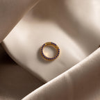 The Pave Amethyst Charm Clip, a gold circular earring adorned with purple gemstones, is displayed on smooth beige satin fabric.