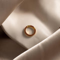 The Pave Garnet Charm Clip, featuring gold and red gemstones, rests on folded champagne satin—ideal for personalizing your jewelry.