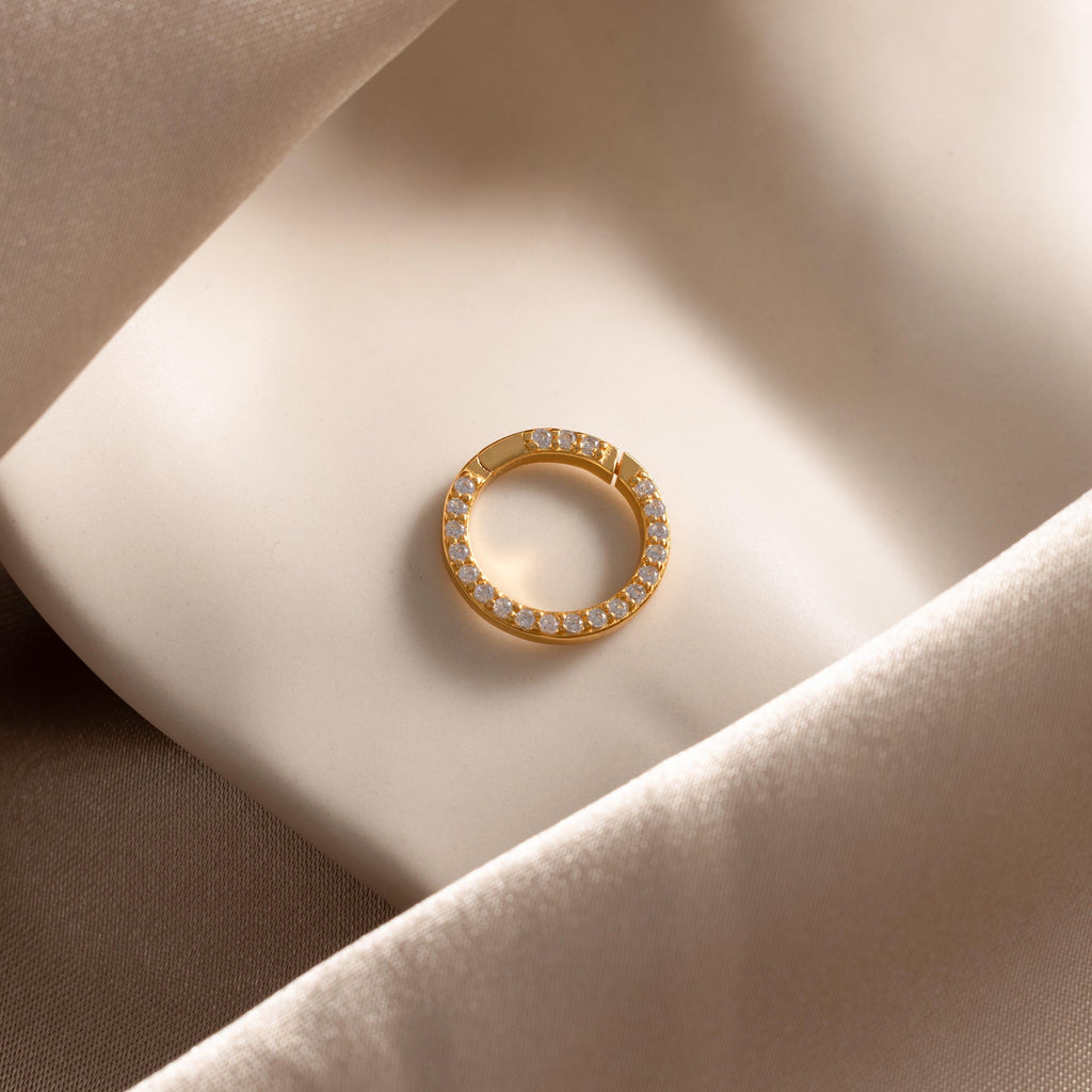 The Pave Diamond Charm Clip, featuring sparkling diamonds, rests on smooth cream-colored satin—ideal for pairing with a personalized jewelry charm holder or a delicate birthstone ring charm.