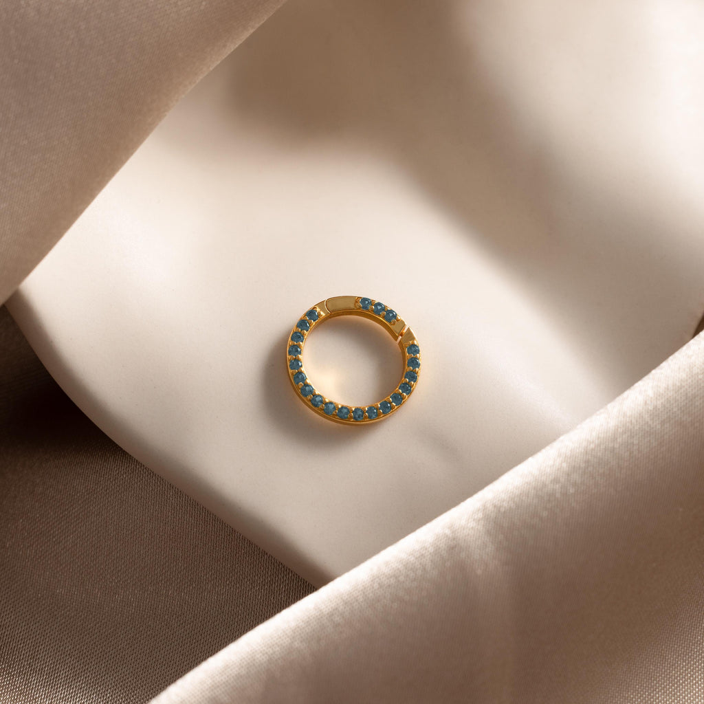 The Pave Blue Zircon Charm Clip, a gold circular earring adorned with blue zircon stones, is displayed elegantly on smooth beige satin fabric.
