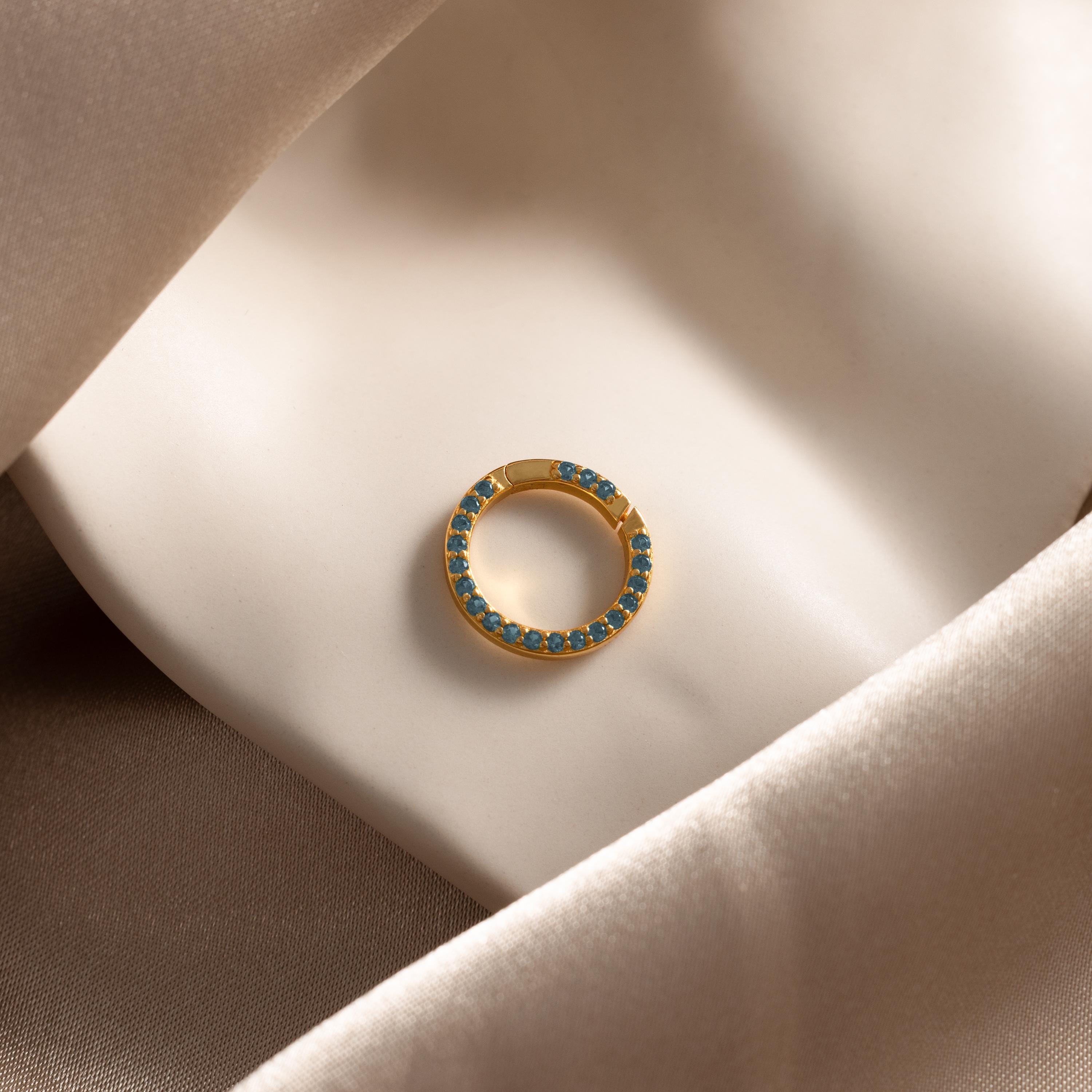 The Pave Blue Zircon Charm Clip, a gold circular earring adorned with blue zircon stones, is displayed elegantly on smooth beige satin fabric.