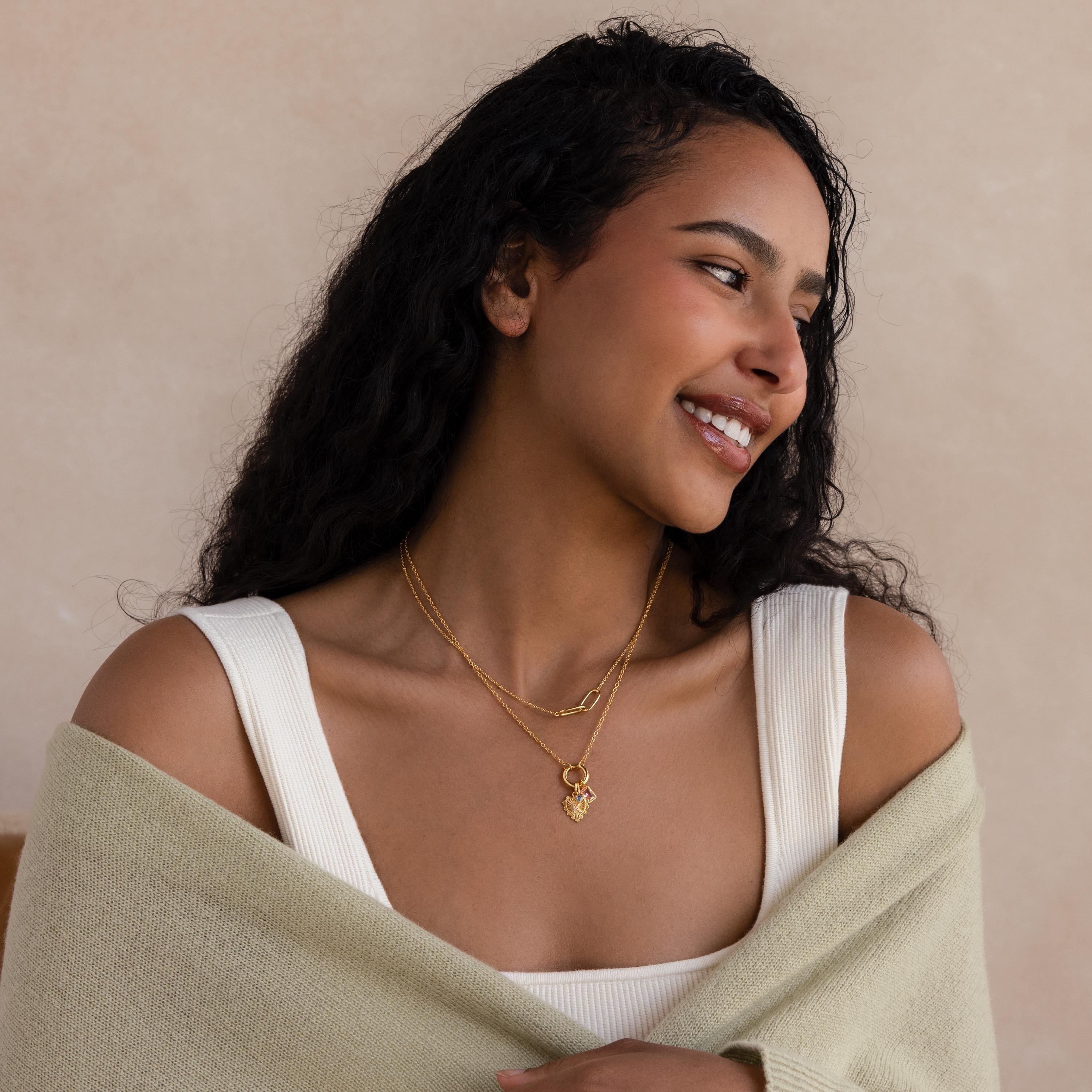 Smiling woman in a white top and light sage shawl, wearing layered gold necklaces including the Cable Circle Charm Necklace in 18K Gold, looking to the side.