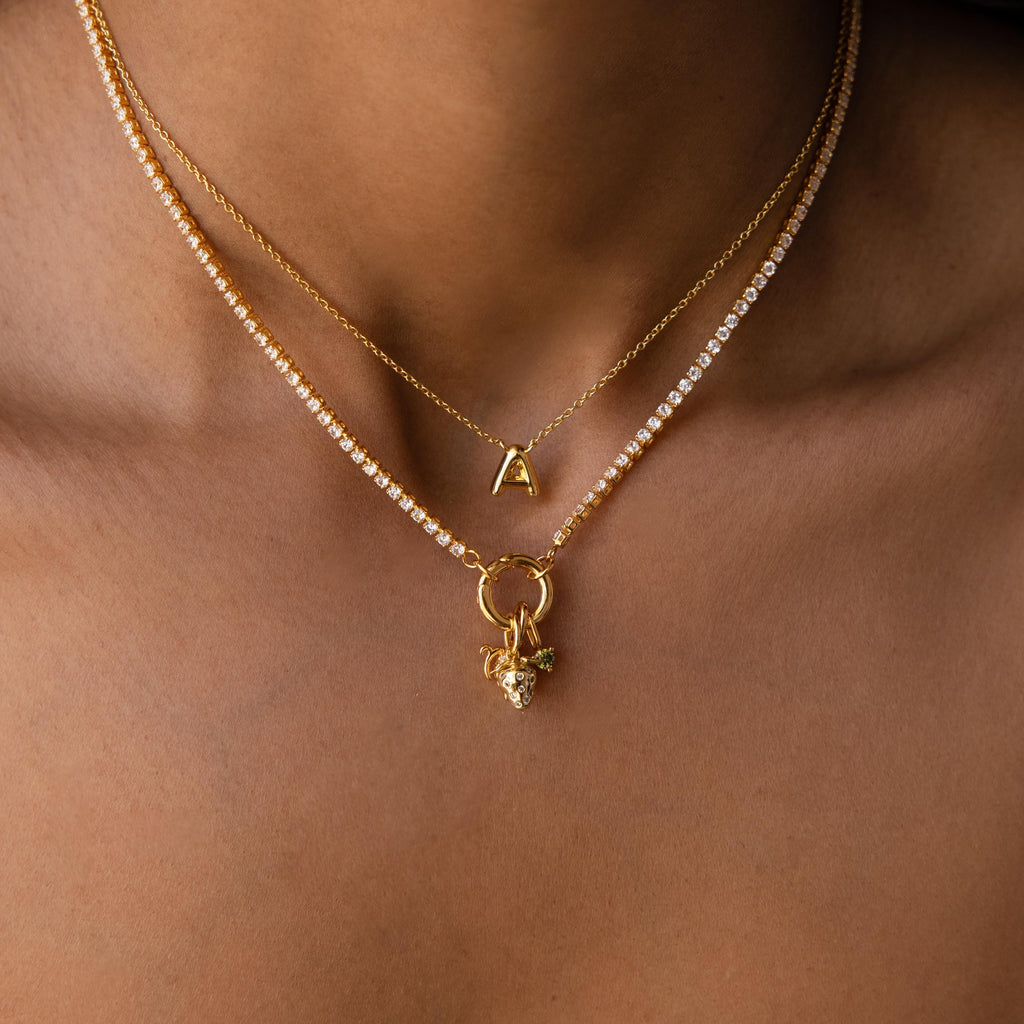 A person wears three gold necklaces: a Diamond Tennis Clasp Necklace, one with a gold letter A pendant, and another with a gold ring and heart-shaped charm secured by a rolo clasp.