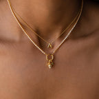 A person wears three gold necklaces: a Diamond Tennis Clasp Necklace, one with a gold letter A pendant, and another with a gold ring and heart-shaped charm secured by a rolo clasp.