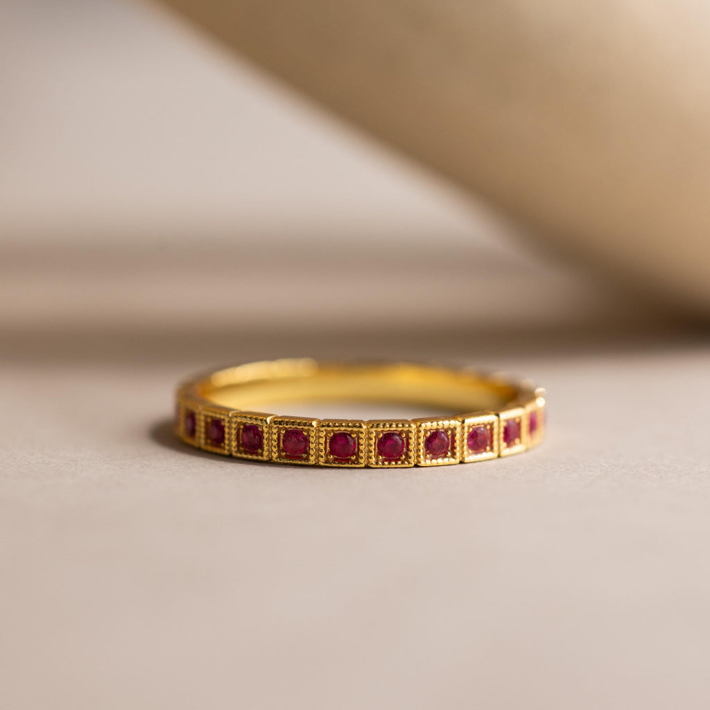 The Art Deco Ruby Eternity Ring features small square rubies set in a vintage milgrain gold bezel, displayed on a neutral surface.