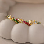 A Duo Initial Peridot Tennis Ring featuring two custom gold initials and sparkling peridot gems rests on a white, sculpted surface resembling teeth.
