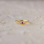 The Amethyst Drop Curve Ring features a delicate gold band with a small amethyst centerpiece, beautifully displayed on a textured light surface. Perfect as a stacking ring for an elegant and subtle accent.