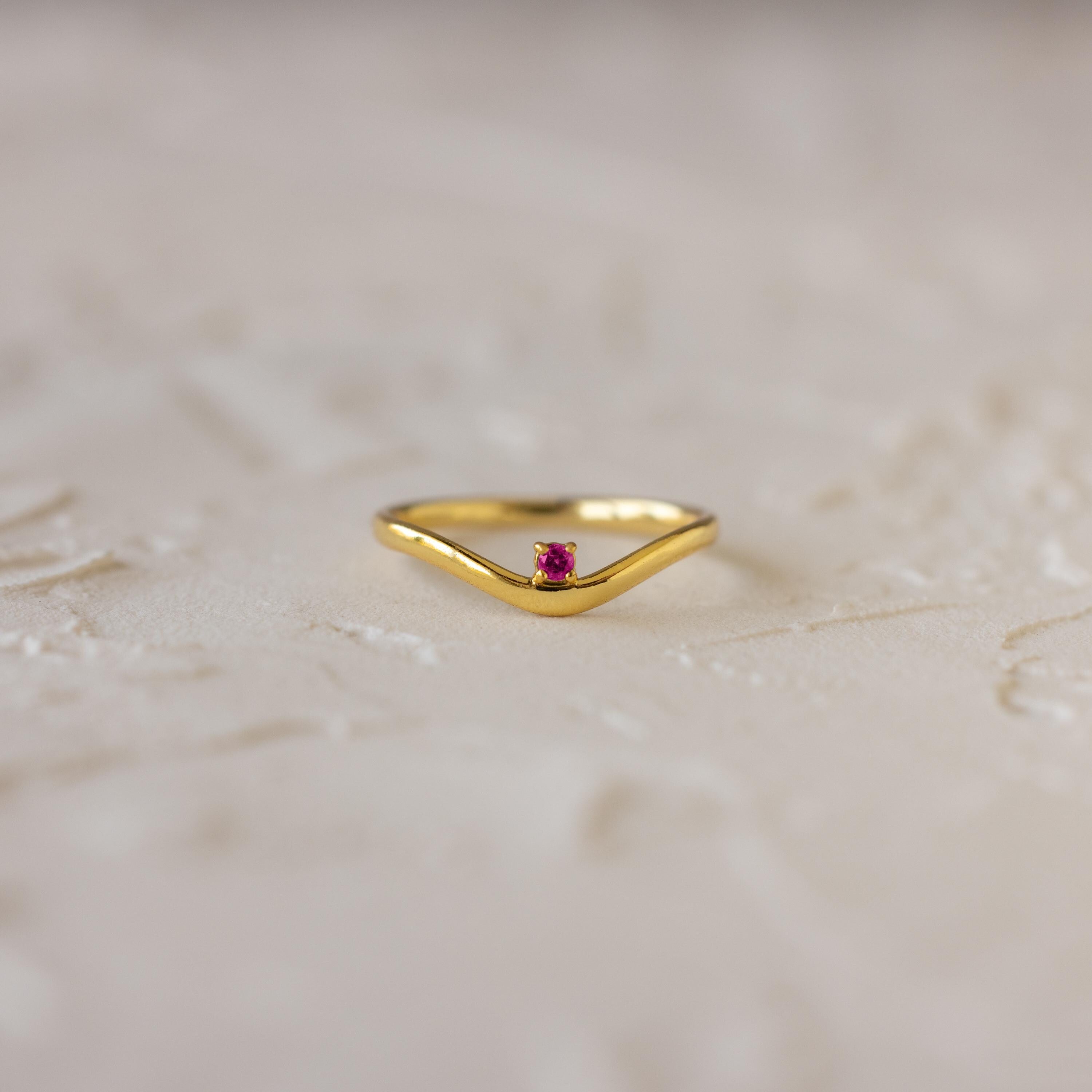 The Amethyst Drop Curve Ring features a delicate gold band with a small amethyst centerpiece, beautifully displayed on a textured light surface. Perfect as a stacking ring for an elegant and subtle accent.