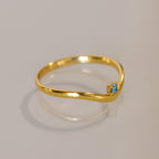 The Blue Zircon Drop Curve Ring features a thin gold band with a small blue zircon gemstone on a gentle curve, ideal as a delicate stacking ring and shown on a light surface.