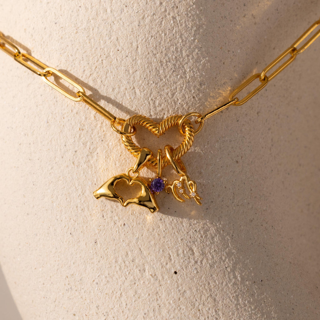 Close up of gold paperclip chain necklace featuring a textured heart-shaped connector that holds three dangling charms: a gold Heart Hands charm with a cut-out heart center, an amethyst birthstone charm, and a zodiac charm