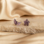 Close up of a pair of purple amethyst moon and star-shaped earrings in silver resting on a beige stone surface.
