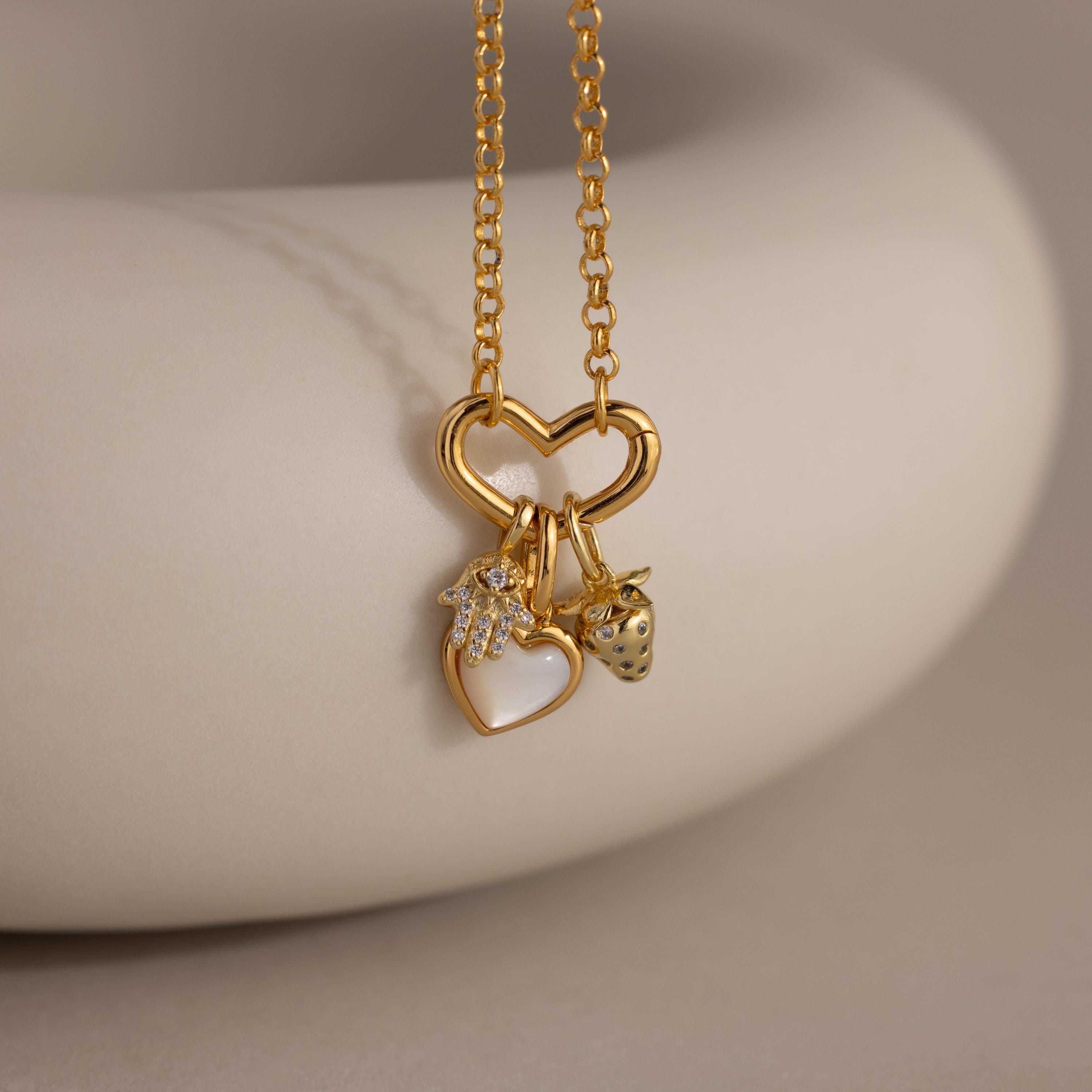 A close-up of a necklace with a gold heart-shaped connector and three charms: a pavé hamsa hand, a mother-of-pearl heart, and a gold strawberry charm, resting on a soft cream ceramic surface.