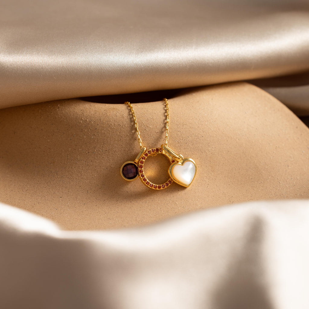 The Pave Garnet Charm Clip—a gold necklace with three charms: a small dark circle, a birthstone ring charm, and a white heart—displayed on satin fabric for an elegant, personalized jewelry look.