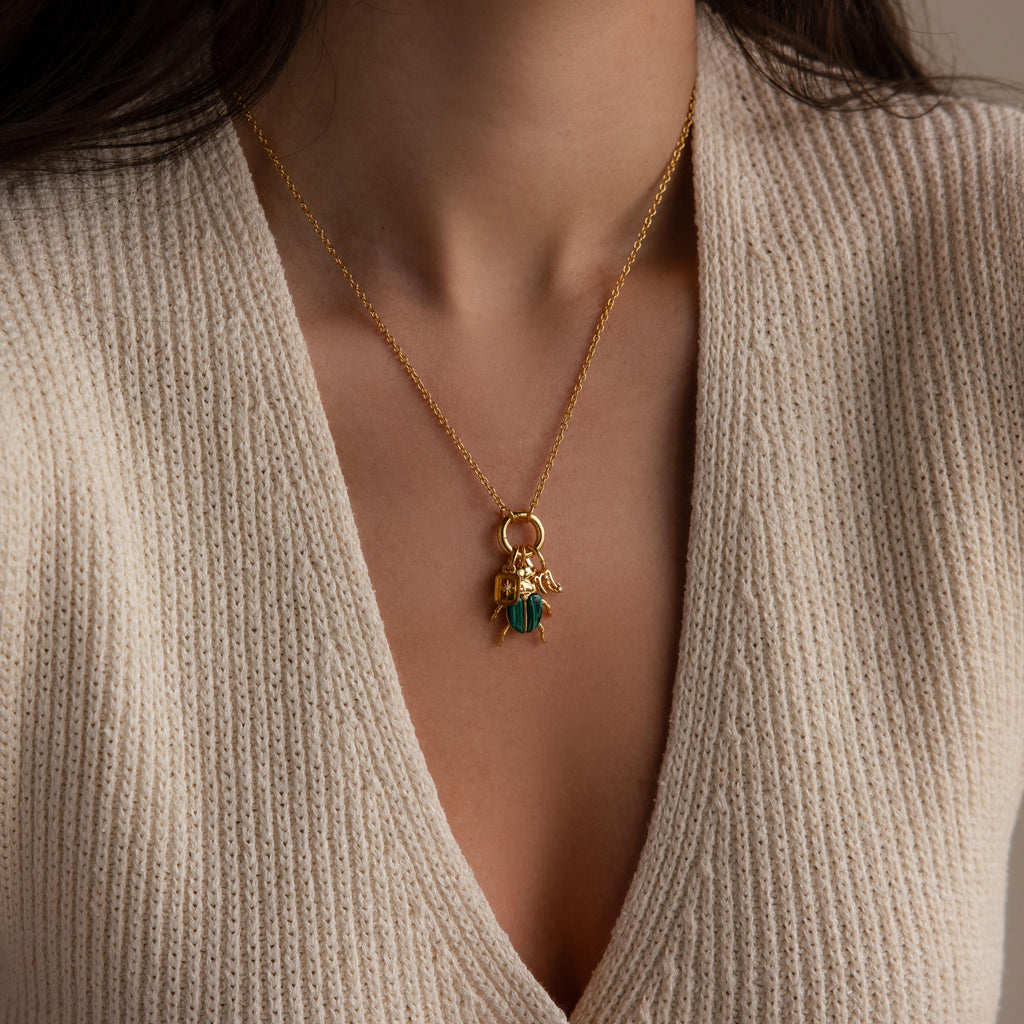 A full view of the beetle charm necklace worn by a model over a beige knit V-neck top. The charm hangs from a gold Rolo Circle Necklace with additional gold charms layered beside it.