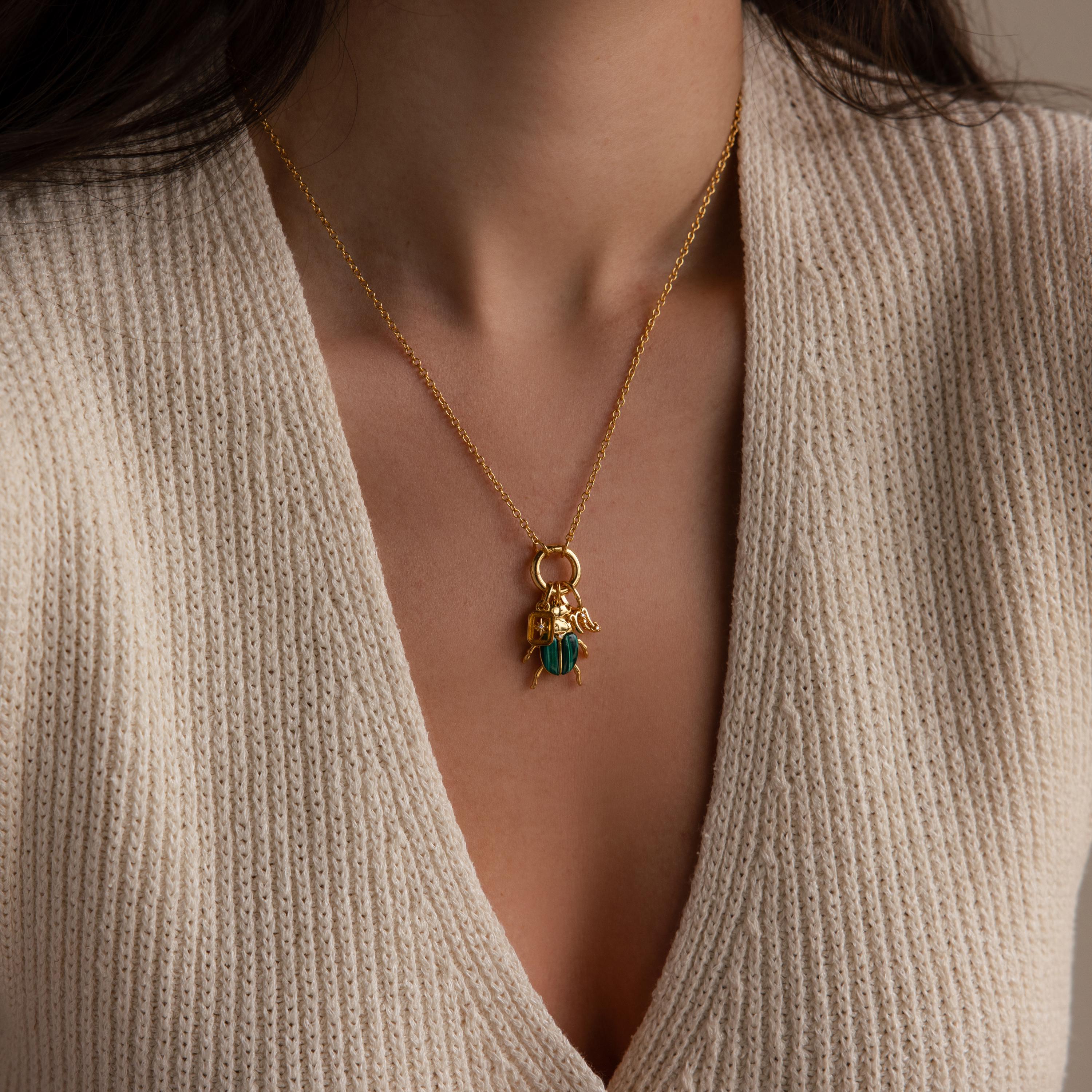 A full view of the beetle charm necklace worn by a model over a beige knit V-neck top. The charm hangs from a gold Rolo Circle Necklace with additional gold charms layered beside it.