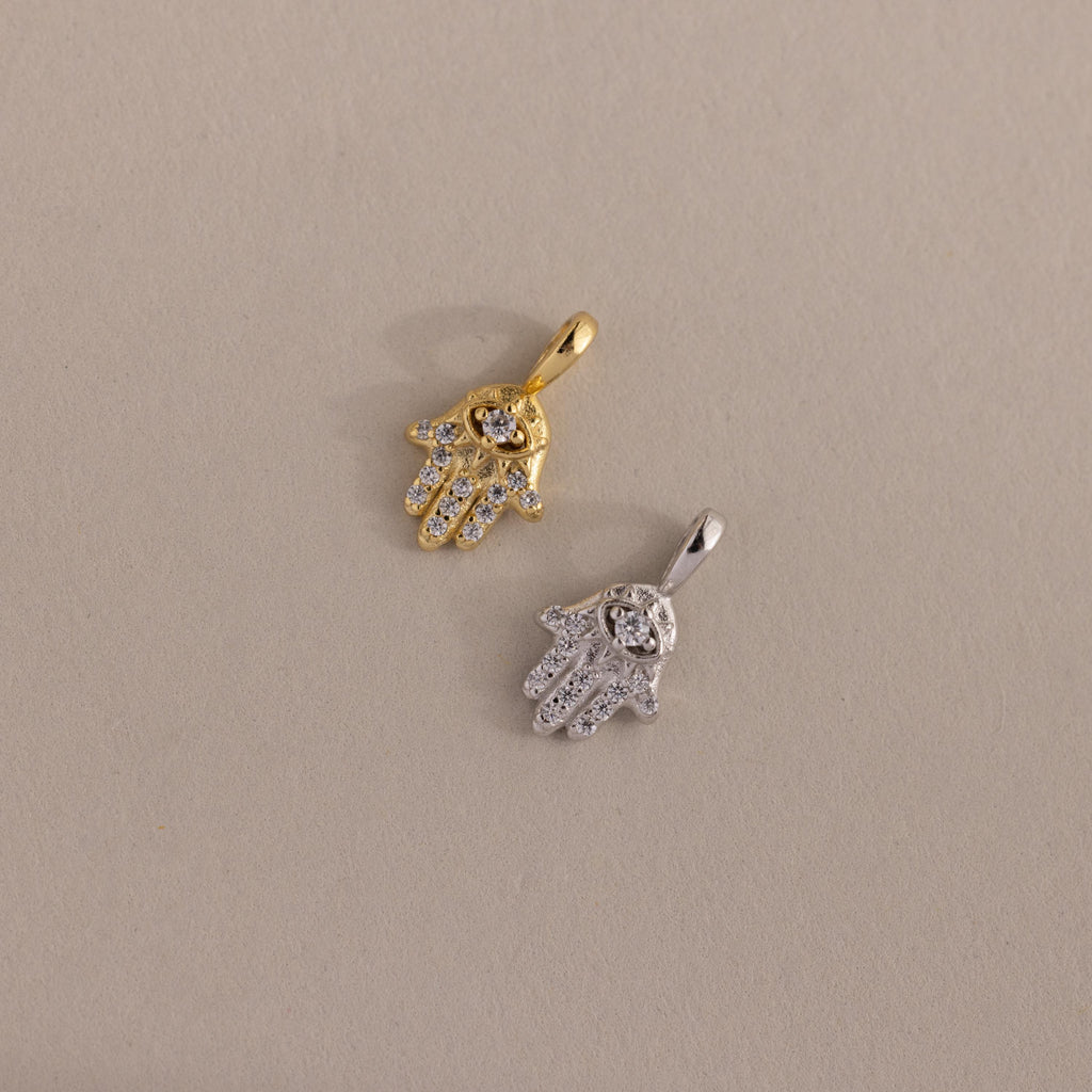 Two Pave Hamsa Hand Charms—one gold, one silver—adorned with clear stones, rest on a beige surface. These charms are perfect protection jewelry for anyone seeking spiritual clarity.