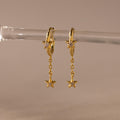 Close of Gold Star Drop Chain Huggie Earrings hanging from a glass.