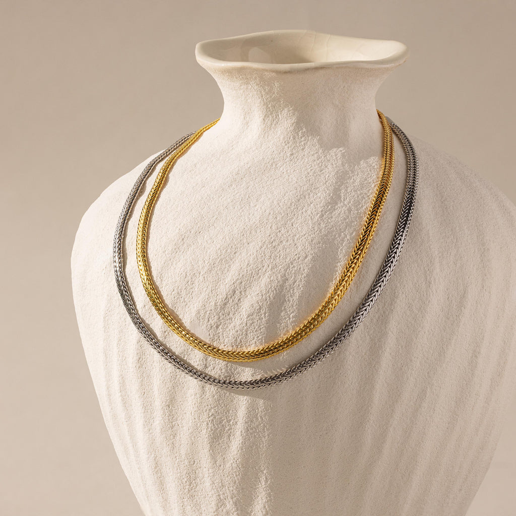 A white textured vase displays a layered necklace with gold and silver woven chains and a Thick Mesh Chain Necklace in White Gold against a neutral background.