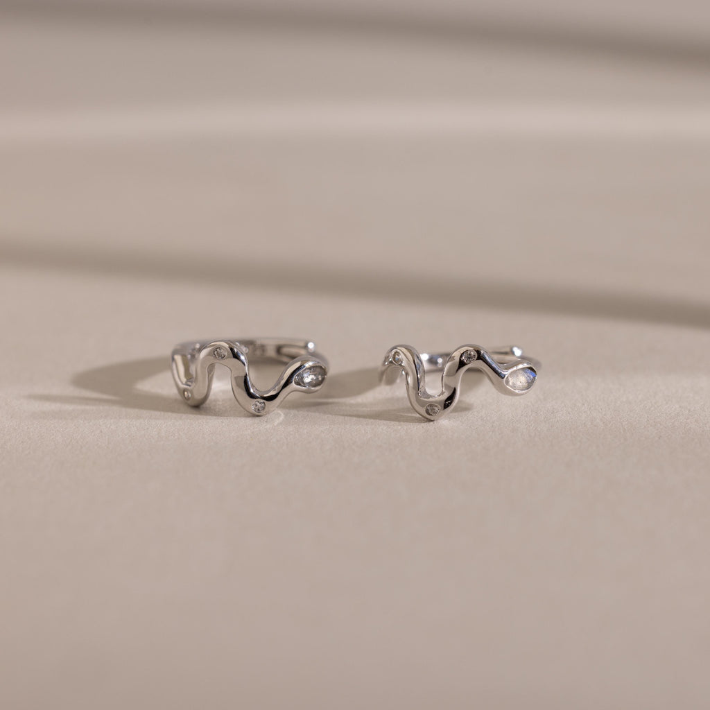 Two wavy, silver Pave Viper Huggies are displayed on a smooth beige surface with soft shadows, highlighting their elegant serpentine curves.