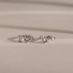 Two wavy, silver Pave Viper Huggies are displayed on a smooth beige surface with soft shadows, highlighting their elegant serpentine curves.