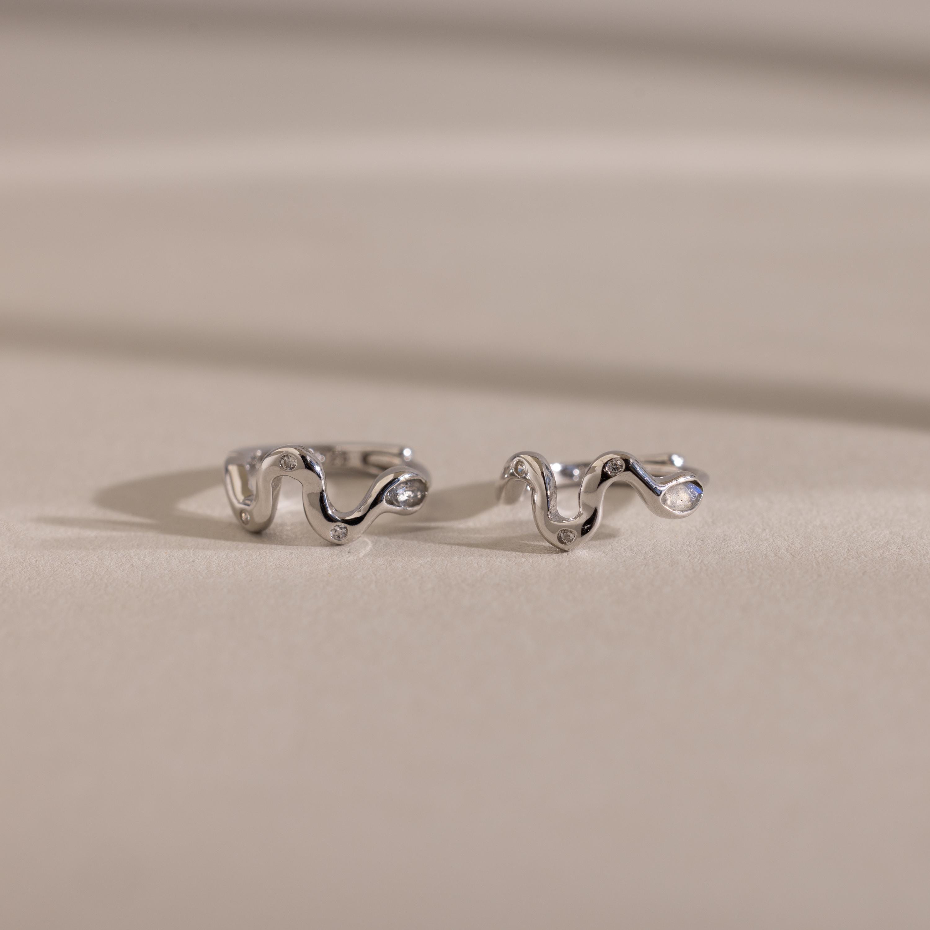 Two wavy, silver Pave Viper Huggies are displayed on a smooth beige surface with soft shadows, highlighting their elegant serpentine curves.