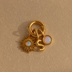 Close-up of three gold charms—an Opal Sun Charm, a bubble letter “S” charm, and a round opal dot charm—hanging together on a polished Rolo Circle connector, displayed on a beige textured surface.