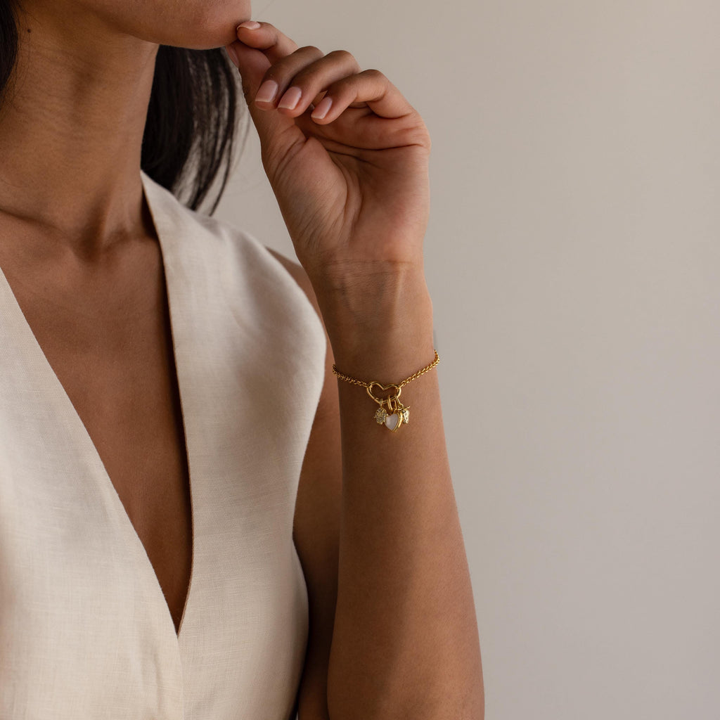 A woman in a sleeveless cream dress wears the Pave Hamsa Hand Charm bracelet, a symbol of spiritual clarity and protection, with her hand resting near her face.