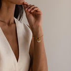 A woman in a sleeveless cream dress wears the Pave Hamsa Hand Charm bracelet, a symbol of spiritual clarity and protection, with her hand resting near her face.