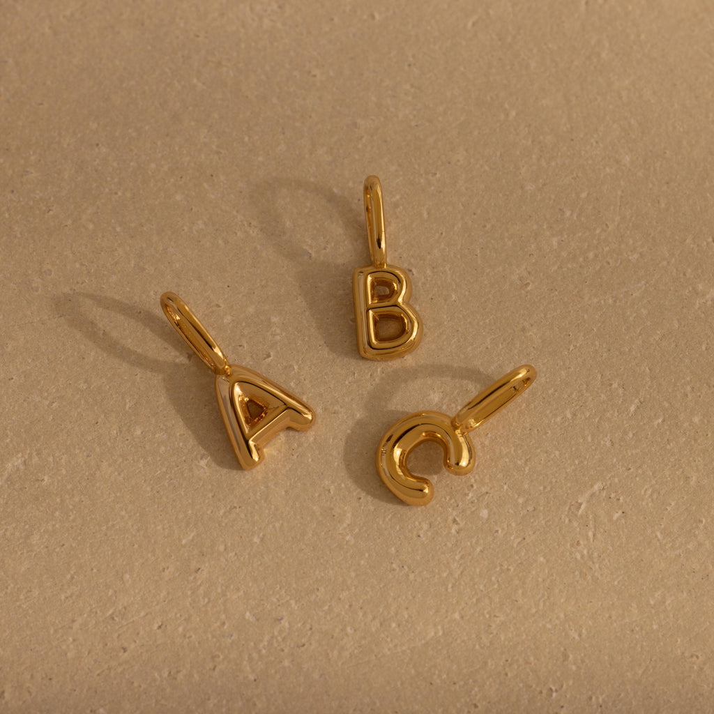 Three bubble letter charms—“A,” “B,” and “C”—in polished gold, arranged in a triangle on a flat beige surface, each featuring a soft, inflated shape and a loop for attachment.