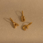 Three bubble letter charms—“A,” “B,” and “C”—in polished gold, arranged in a triangle on a flat beige surface, each featuring a soft, inflated shape and a loop for attachment.