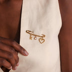 Gold safety pin with beaded accents pinned on a v-neck cream garment, styled with a pearl, gold heart, and initial K charm.