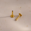 A pair of 18k gold Flat Back Posts positioned on a speckled ceramic surface, one standing and one lying down.
