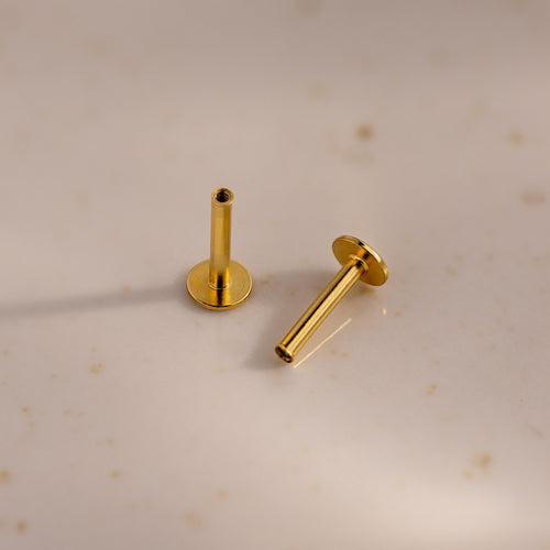 A pair of 18k gold Flat Back Posts positioned on a speckled ceramic surface, one standing and one lying down.
