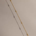 Side-by-side flat-lay of the Dainty Bow Necklace in both gold and silver finishes on a soft beige background.