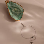 Flat lay of a dainty silver hand chain bracelet displayed on a soft beige surface, with the chain draped across a green and gold oyster shell. The bracelet features three bezel-set diamonds and a heart charm at the lobster clasp.