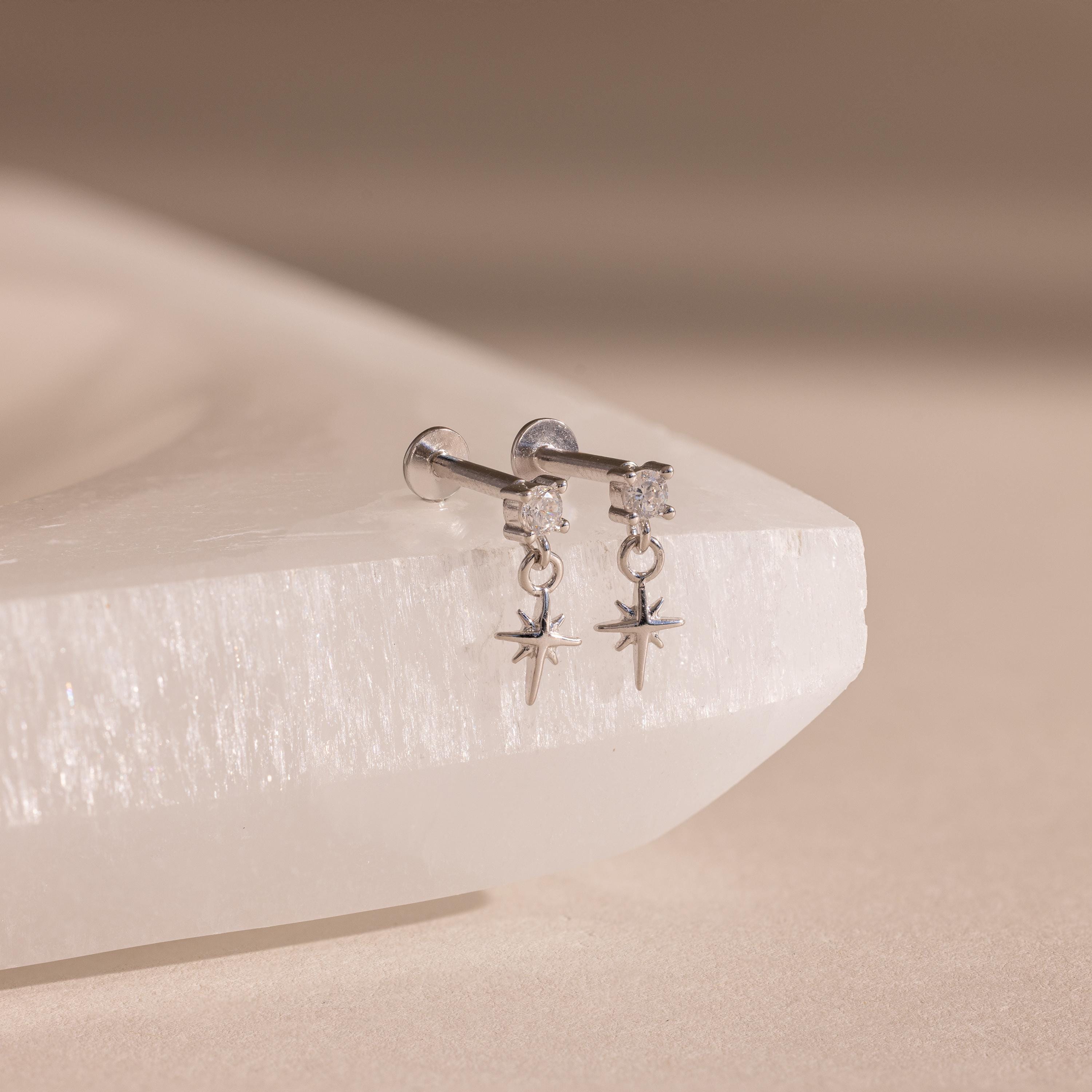 Pair of silver flatback earrings featuring a small round diamond stud and dangling star charms, displayed on a textured white crystal surface.