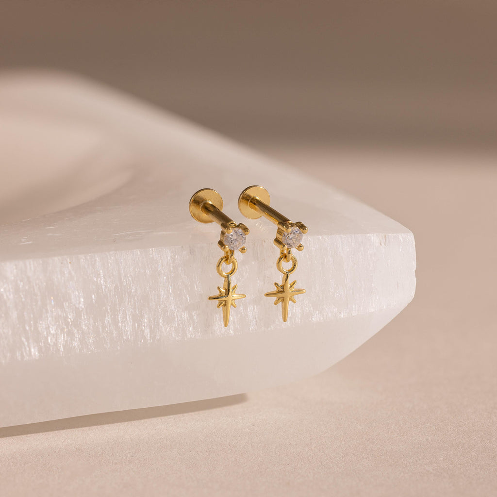 A close-up product shot of the Diamond Star Flatback Earrings in an 18k gold finish. Each earring features a clear, round-cut gemstone set in a prong setting at the top. Hanging delicately from a small gold loop below the stone is a finely crafted star charm, designed with four elongated arms—vertical and horizontal—with shorter, angled rays between them, mimicking a celestial sparkle.