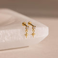 A close-up product shot of the Diamond Star Flatback Earrings in an 18k gold finish. Each earring features a clear, round-cut gemstone set in a prong setting at the top. Hanging delicately from a small gold loop below the stone is a finely crafted star charm, designed with four elongated arms—vertical and horizontal—with shorter, angled rays between them, mimicking a celestial sparkle.