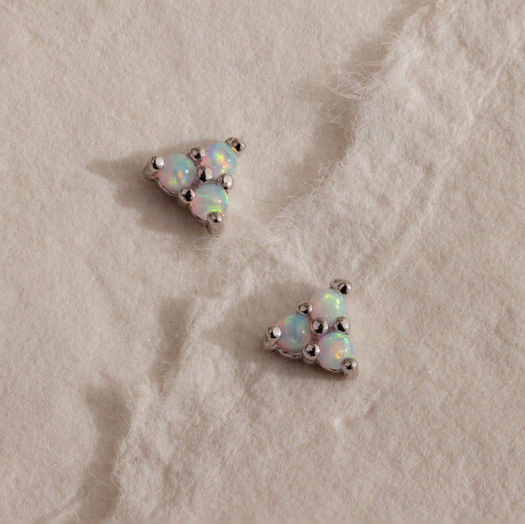 A pair of silver Opal Cluster Stud Earrings resting on a white textured paper, with a triangular trio of luminous white opals set in polished silver.