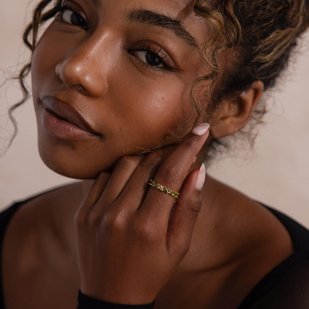 Close up of a woman’s face and hand, wearing a gold ring with marquise cut green ombre stones that shift from pale yellow green to deep olive, styled on her middle finger.