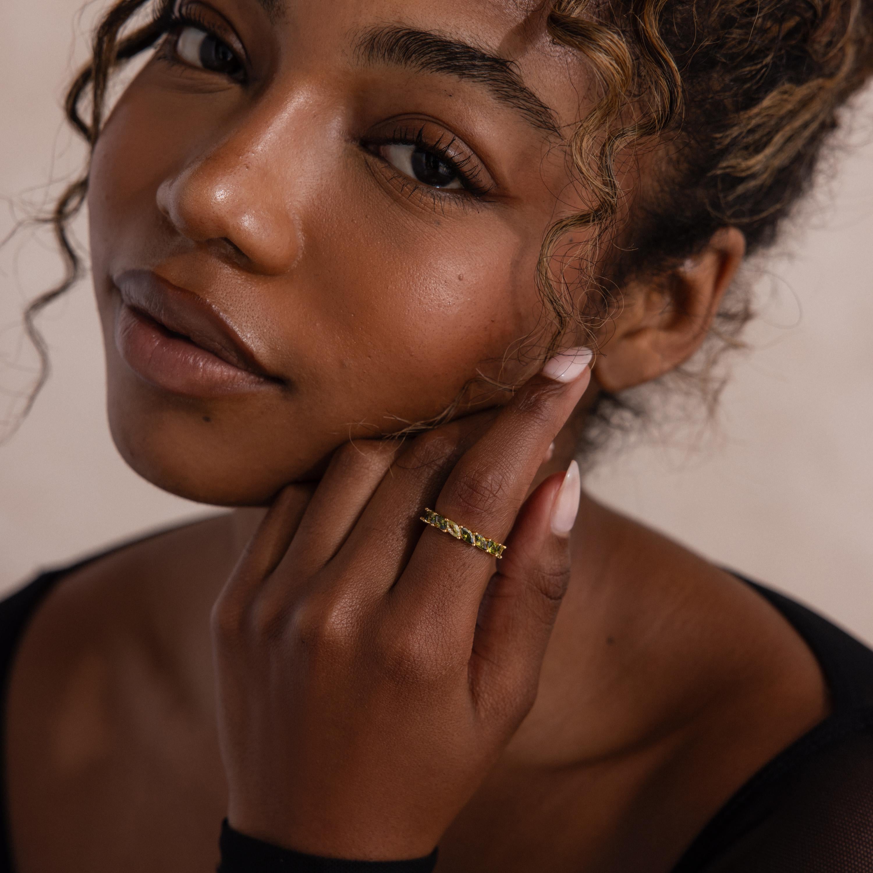 Close up of a woman’s face and hand, wearing a gold ring with marquise cut green ombre stones that shift from pale yellow green to deep olive, styled on her middle finger.