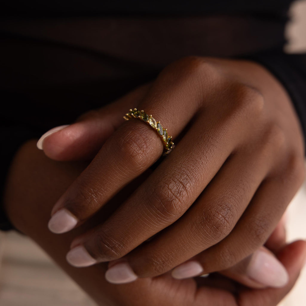 Close up of a hand wearing a gold ring set with marquise cut green ombre stones that shift from light chartreuse to deep olive. The ring stands out against the model’s dark skin tone and soft pink manicure.