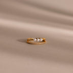 Gold duo ring featuring one band with three round white pearls and a second slim band with pavé crystals, displayed on a smooth beige surface.