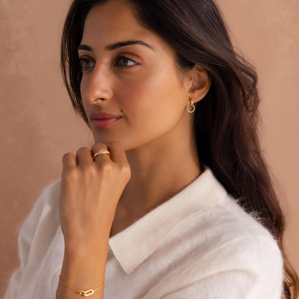 A woman with long dark hair, wearing a white top and elegant Dainty Duo Pave Huggies, featuring interlocking circles, gazes thoughtfully to the side.